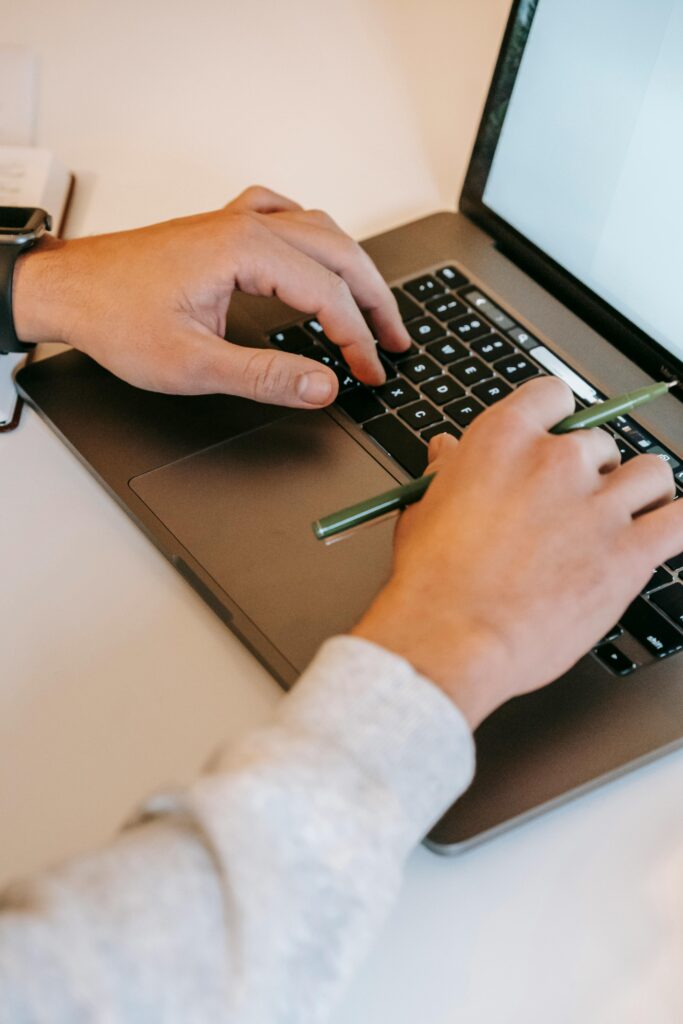 Close-up of hands typing on a laptop keyboard while holding a pen, showcasing productivity.